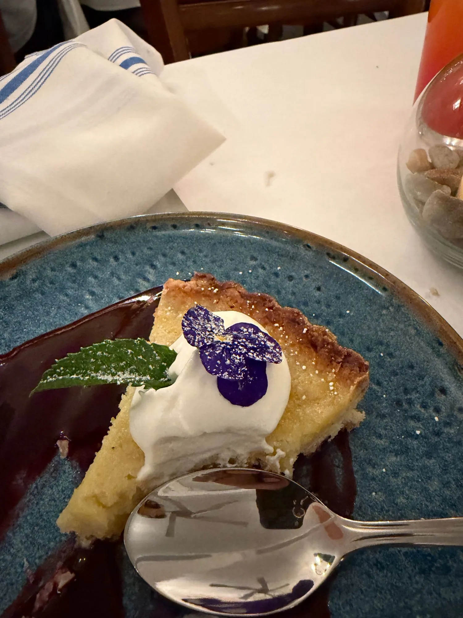 A slice of lemon tart with whipped cream, mint and a violet on a blue plate